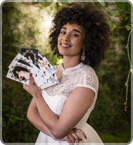 A smiling person with curly hair in a white lace dress holds a fan of portrait cards in a lush, green outdoor setting.