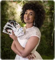 A person with curly hair smiles while holding a fan of small photographs against a lush, green outdoor background.