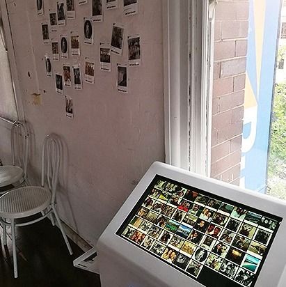 A kiosk displays a digital photo collage next to a wall covered in printed photos and two white metal chairs by a window.