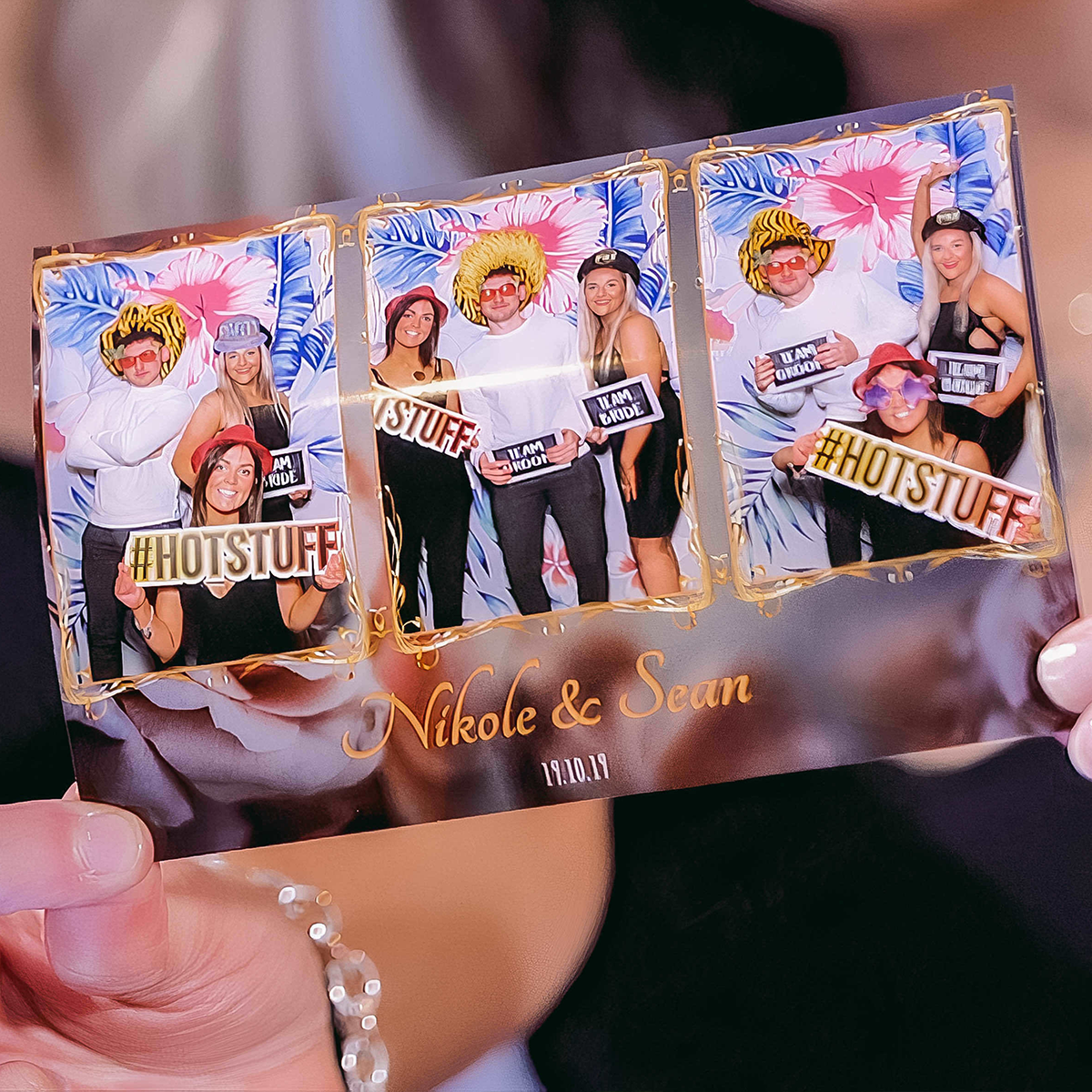 A photo booth strip held by a person, featuring three photos of a group with props and text reading “Nikole & Sean 2023.”