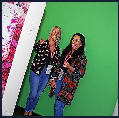 Two people wearing lanyards stand in front of a green screen, one pointing toward a side wall decorated with pink flowers.