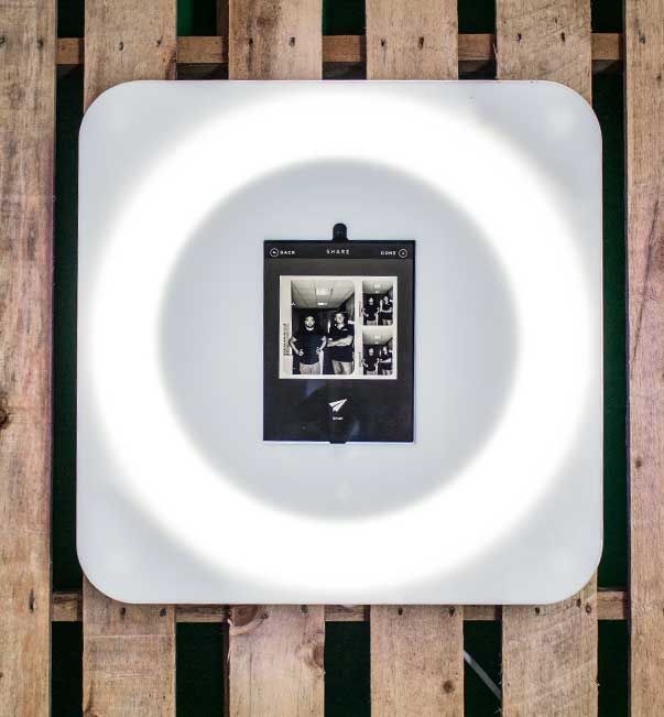 A black-and-white photo booth strip is displayed on a lit square panel against a wooden pallet background.