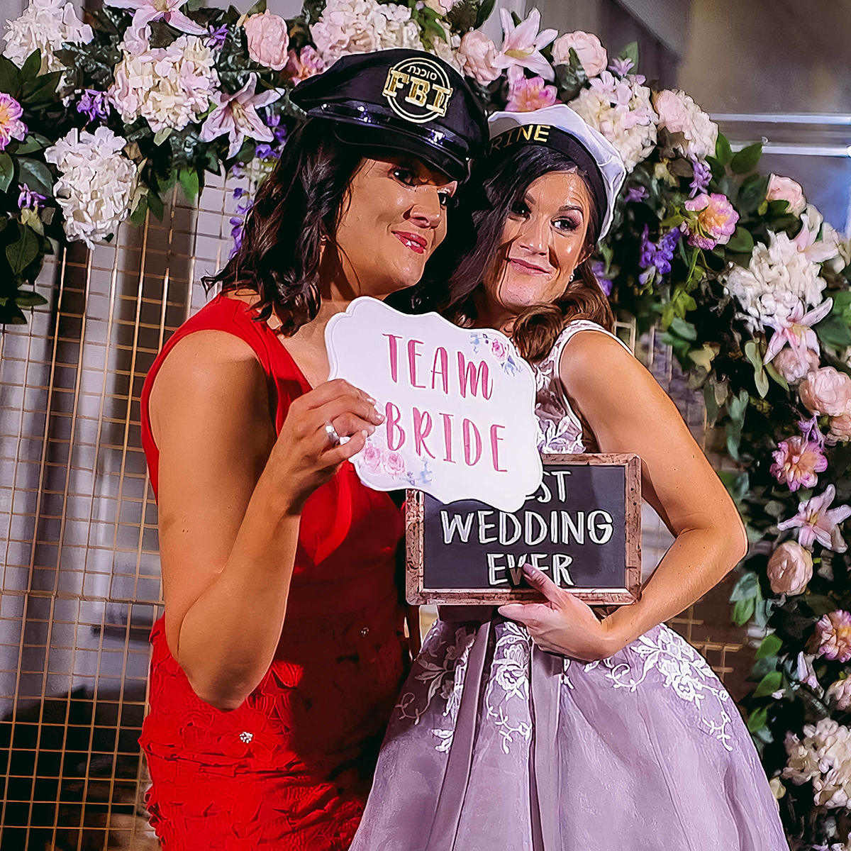 Two people in party hats pose by flowers, holding signs that say