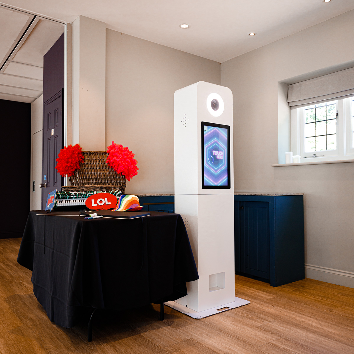 A white photo booth stands beside a table covered in a black cloth with party props in a bright room.