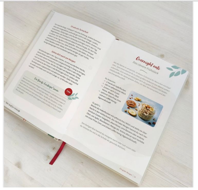 Een open receptenboek ligt op een witte tafel, met twee ge&iuml;llustreerde pagina's waarop tekst en een foto van een gerecht te zien zijn.