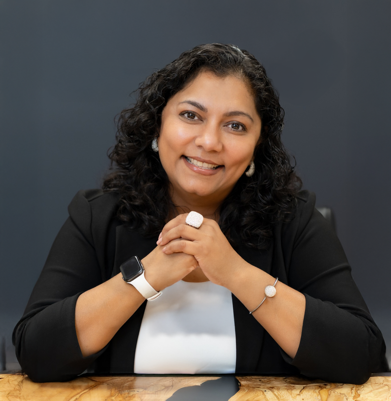 A smiling woman with dark curly hair in a black blazer and white shirt, sitting at a desk with folded hands.