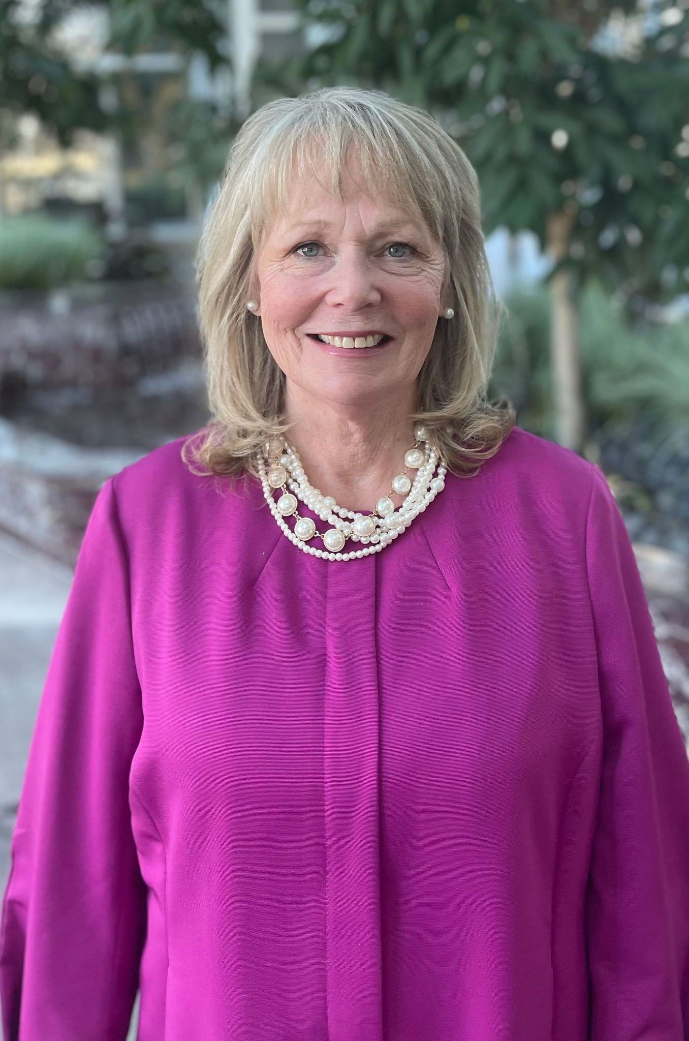 A woman wearing a purple jacket and a pearl necklace is smiling for the camera.
