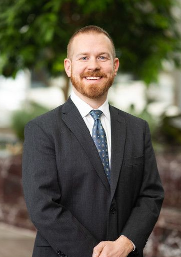 A man in a suit and tie is standing with his hands folded in front of a tree.