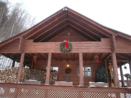 Log cabin with Christmas decorations, porch, and stacked firewood; snow-covered landscape.