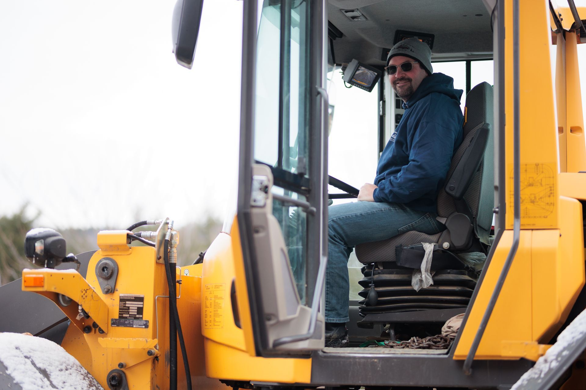 Man in a yellow tractor cab, smiling. Snowy outdoor setting.