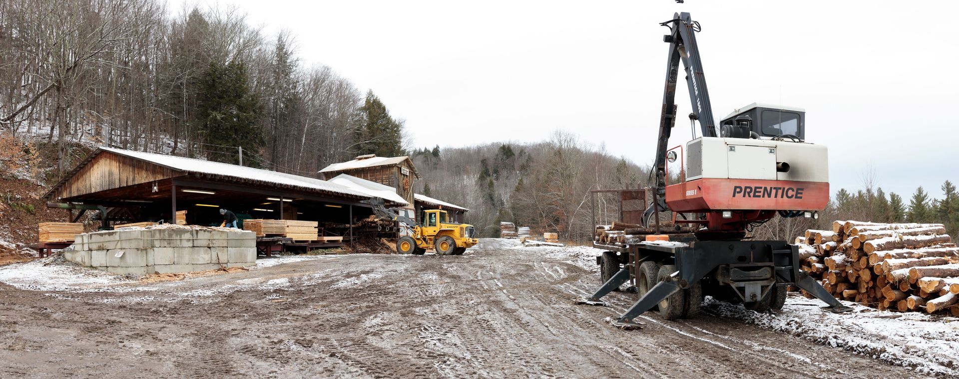 A lumber yard with a crane and wood logs on a snowy day.