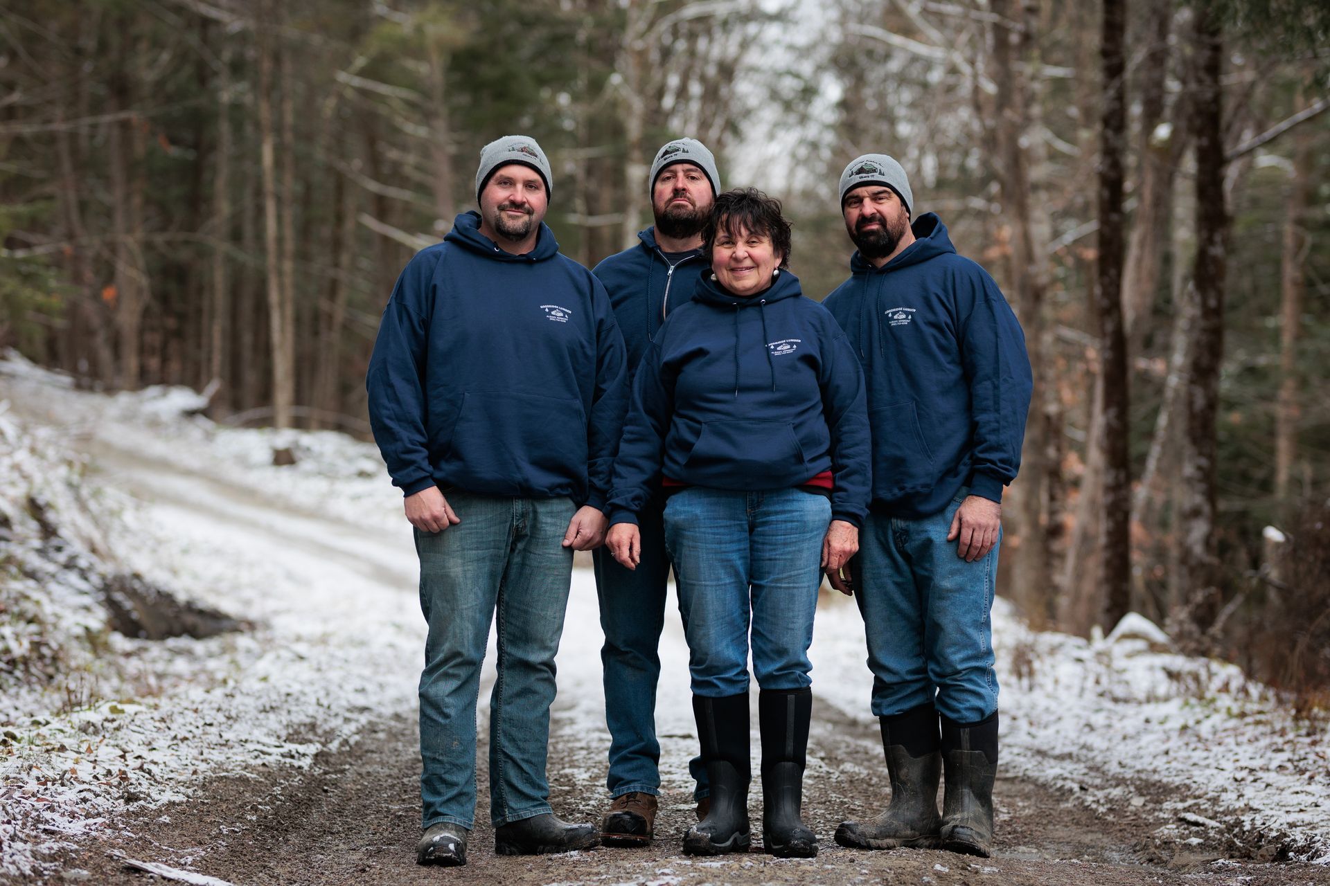 Four people wearing matching blue sweatshirts and knit caps stand on a snowy dirt road in a forest.