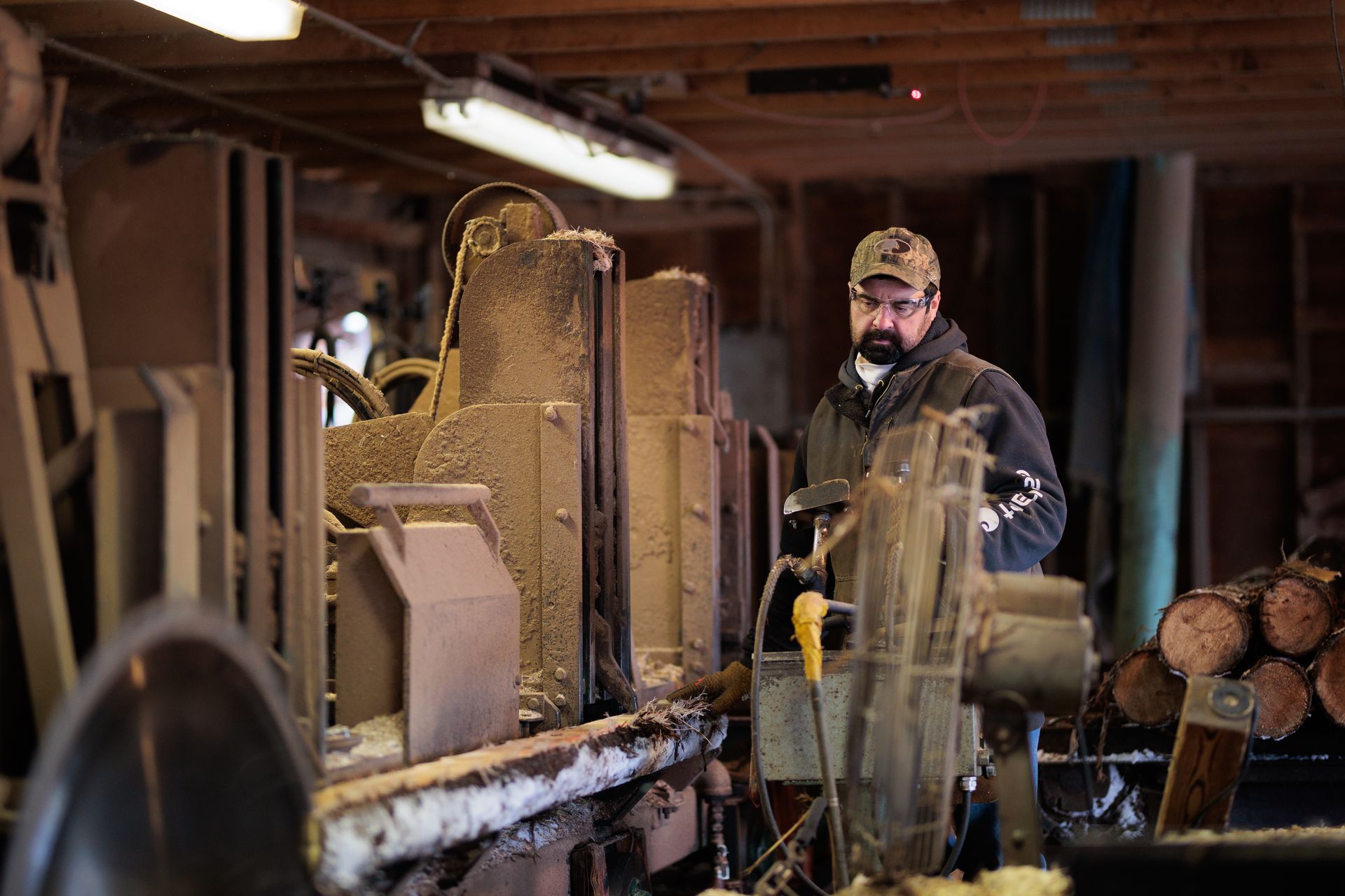 Man operating a sawmill in a wood-filled workshop. He's wearing a camo cap, standing near machinery.