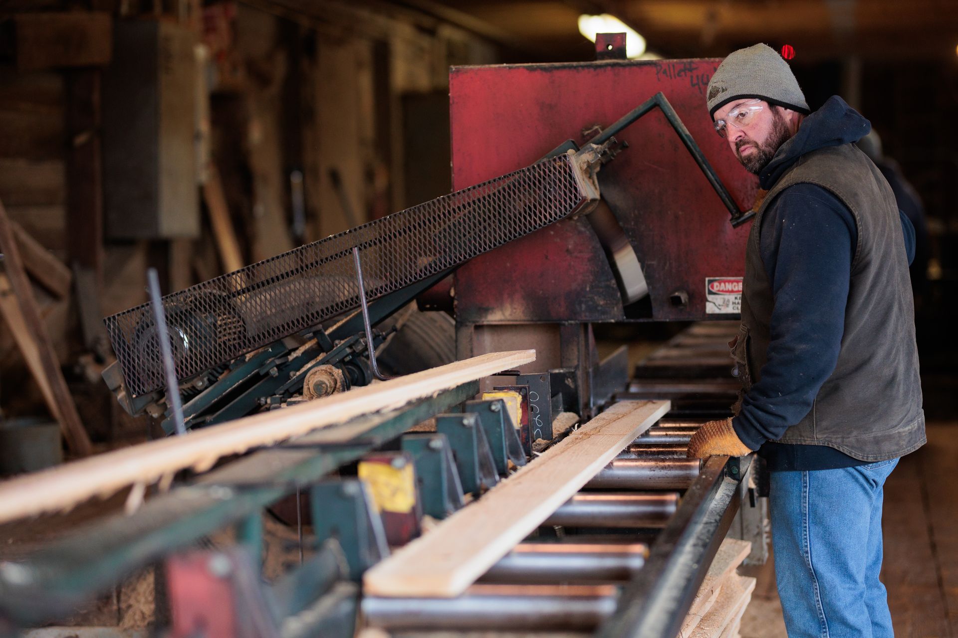 Man in a beanie and vest watches lumber moving through a sawmill.