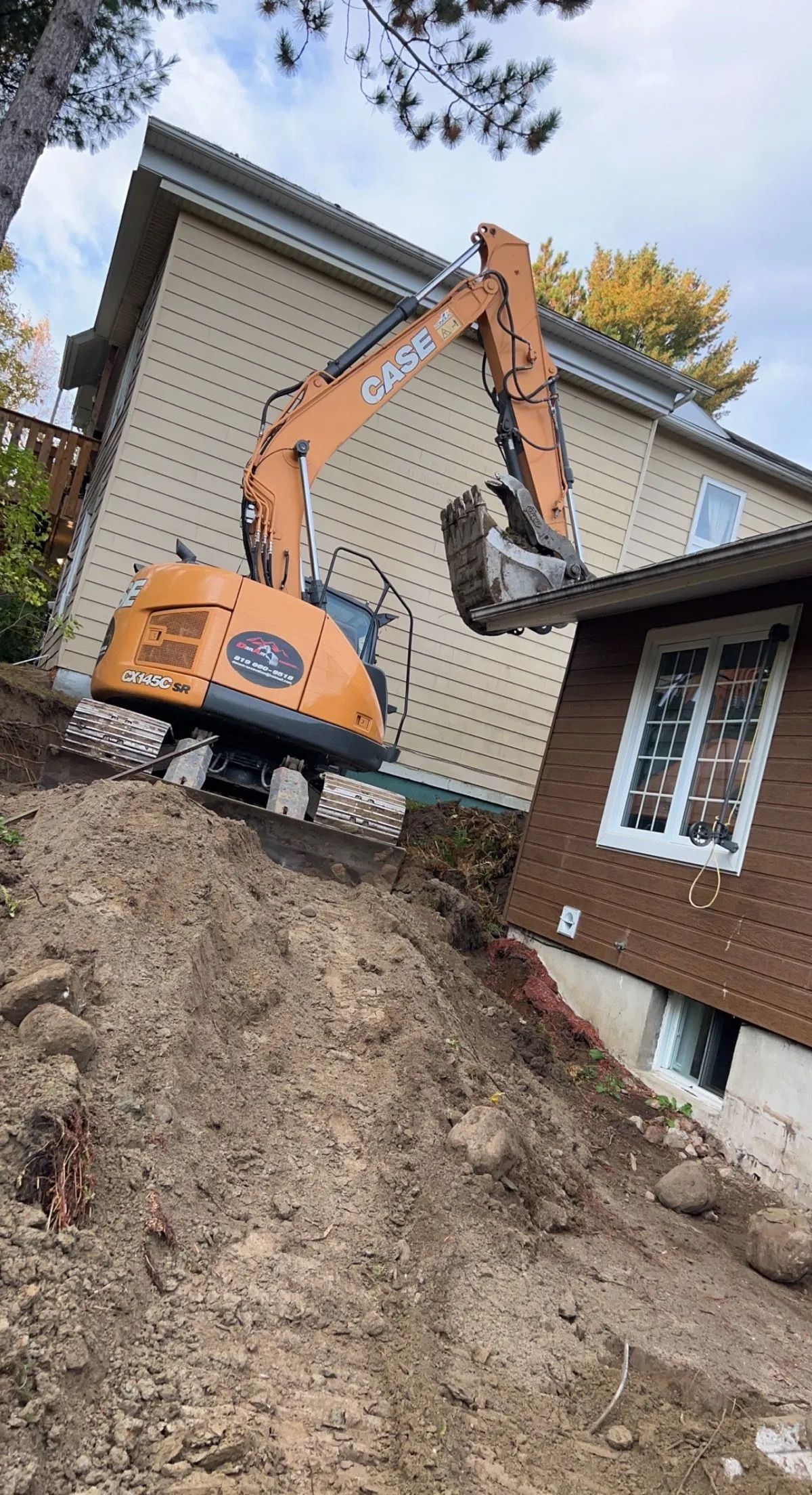 Une excavatrice Case orange est posée sur un terrain en pente et en terre battue, à côté d'une maison brune, son bras étant levé près de la ligne de toit.