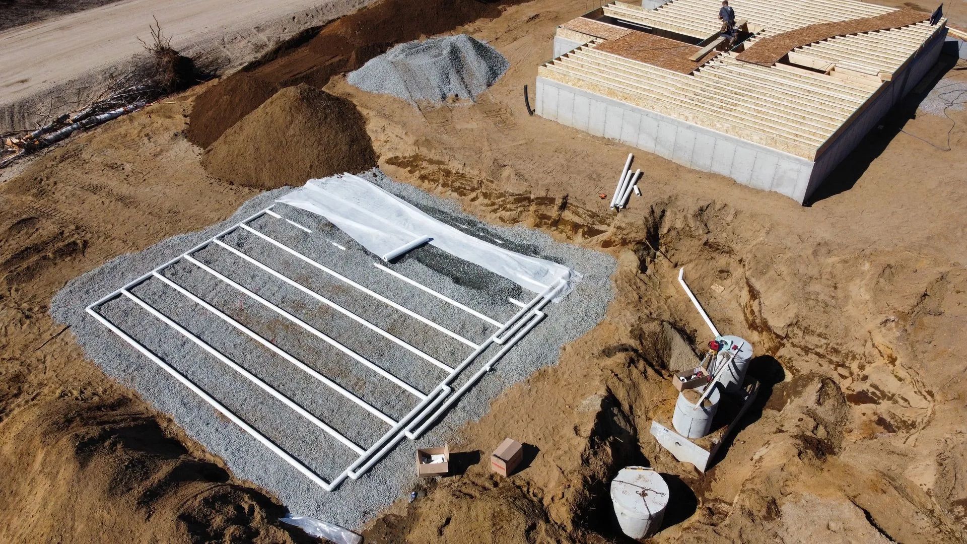 Vue aérienne d'un chantier de construction montrant un champ d'épandage septique rempli de gravier avec des tuyaux blancs à côté d'un bâtiment.