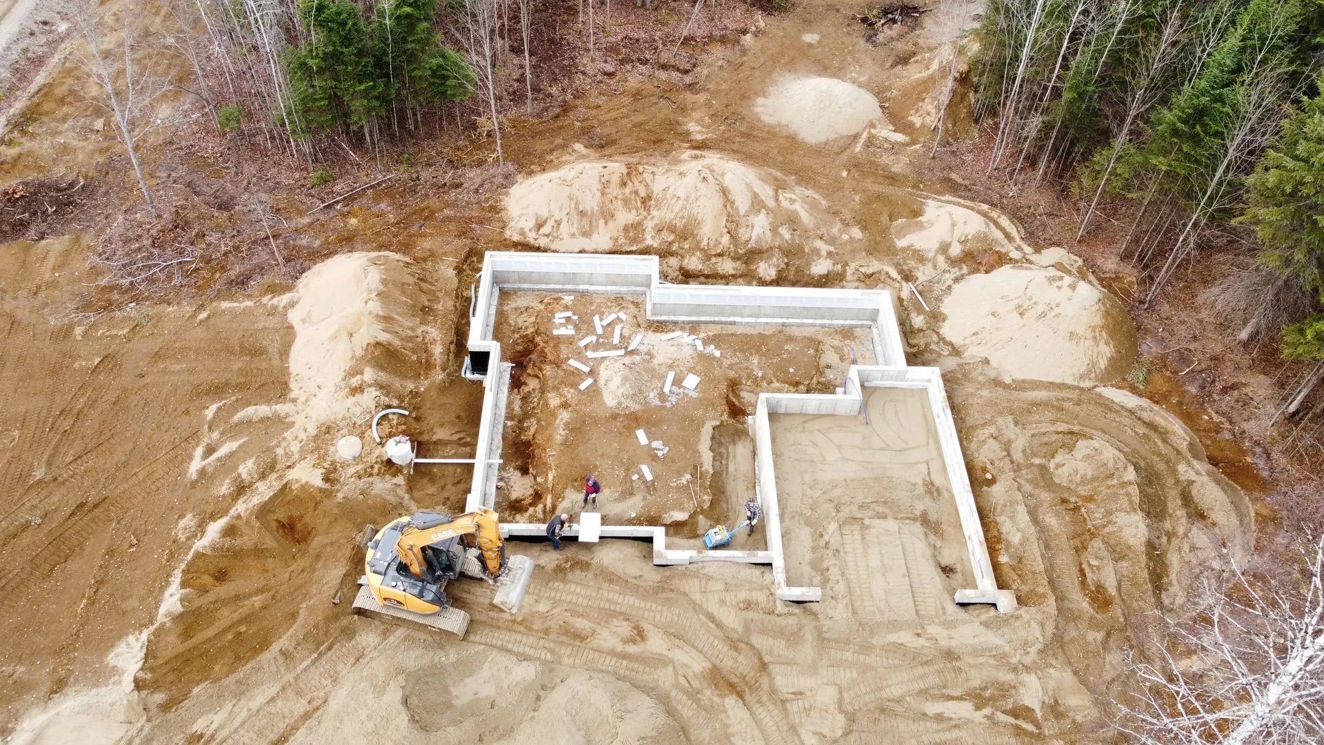 Vue aérienne d'un chantier de construction montrant les fondations d'une maison en béton avec une excavatrice garée à proximité.