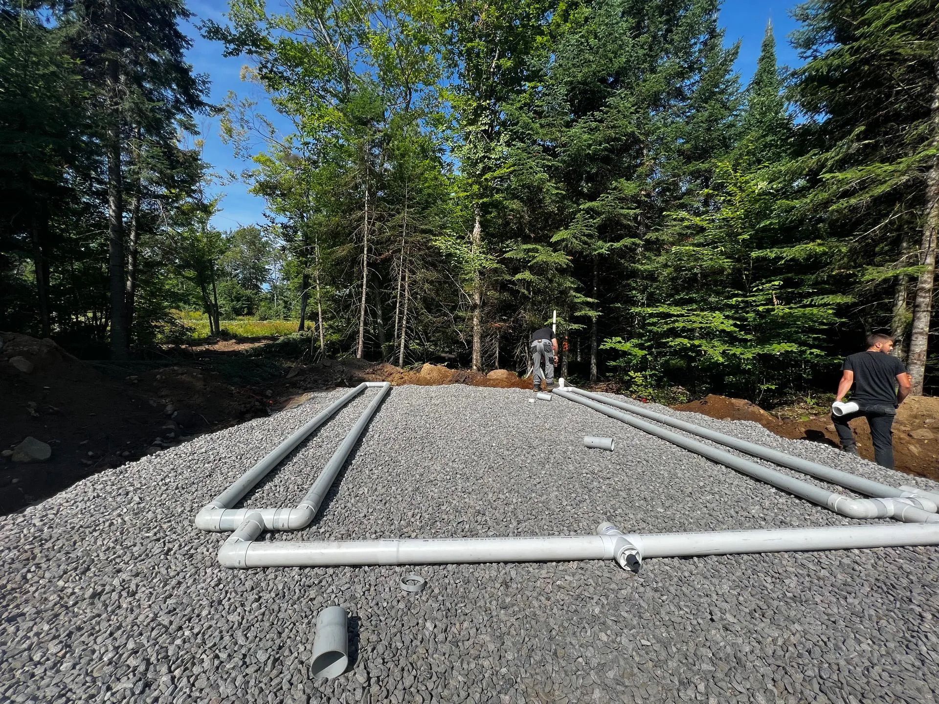 Un lit de gravier rectangulaire avec un réseau de tuyaux en PVC blanc installé dans une zone boisée, avec deux personnes travaillant à l'arrière.