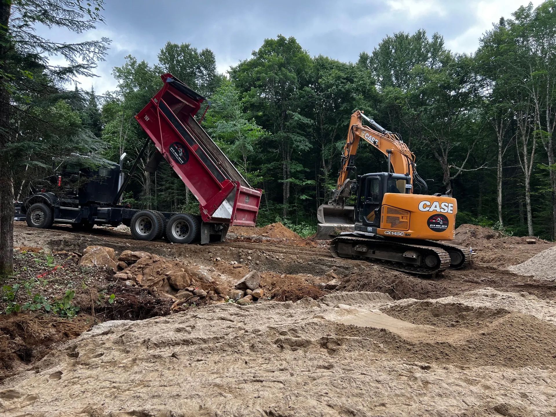 Un camion-benne rouge, benne levée, et une pelle mécanique Case jaune travaillent sur un chantier de construction en terre battue dans les bois.