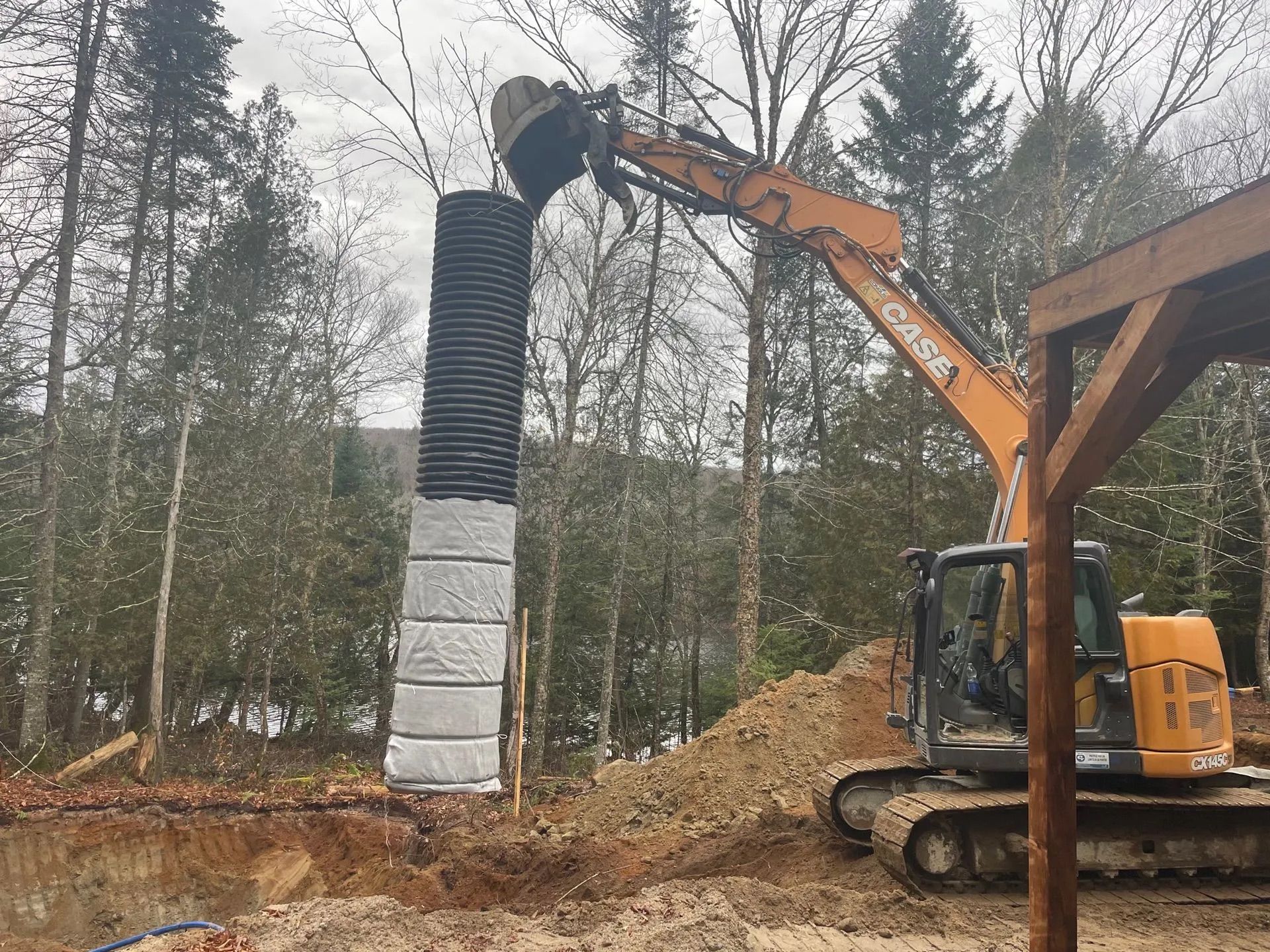 Une excavatrice soulève un tuyau de drainage vertical noir et blanc ondulé au-dessus d'un chantier de construction extérieur.