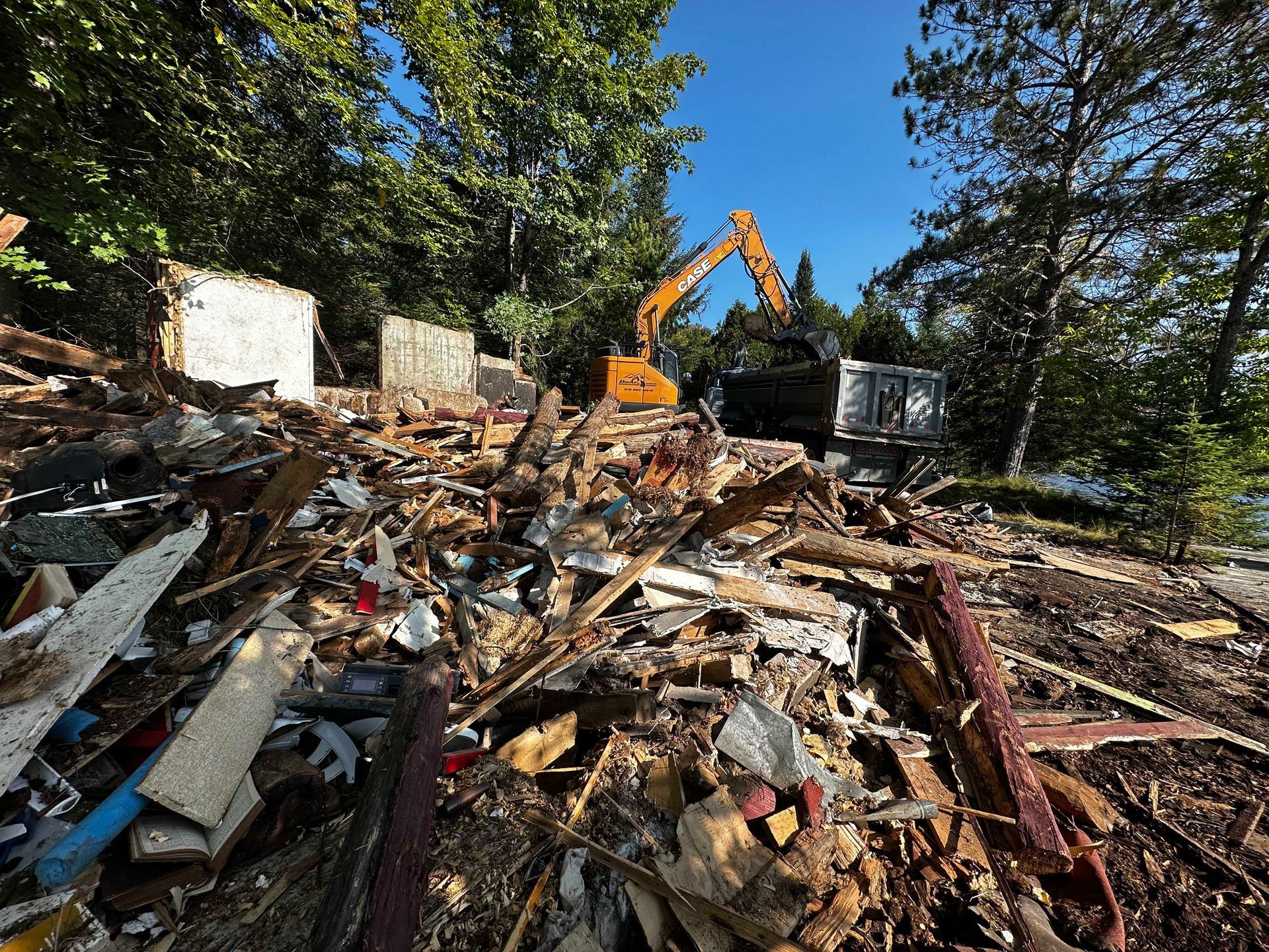 Une pelleteuse orange dégage les débris d'un chantier de démolition dans une zone boisée, sous un ciel bleu azur.