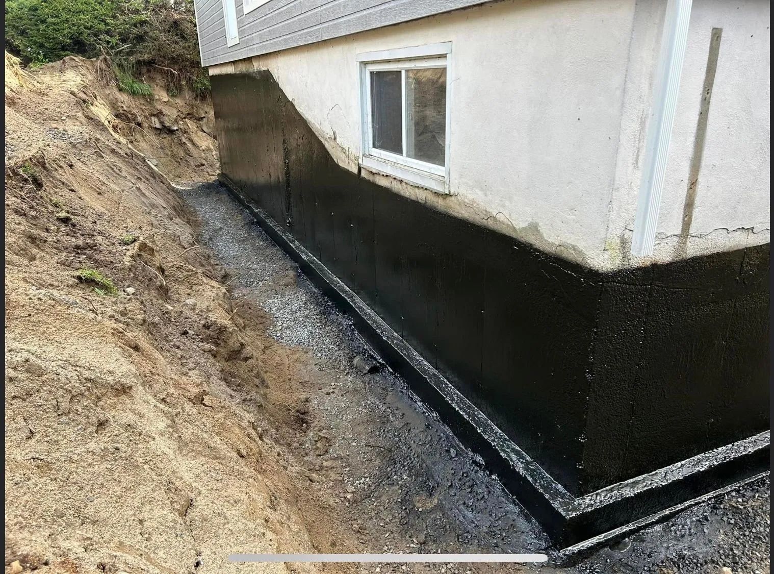 Mur de fondation fraîchement recouvert d'un mastic d'étanchéité noir, avec une tranchée remplie de gravier le long du bâtiment.