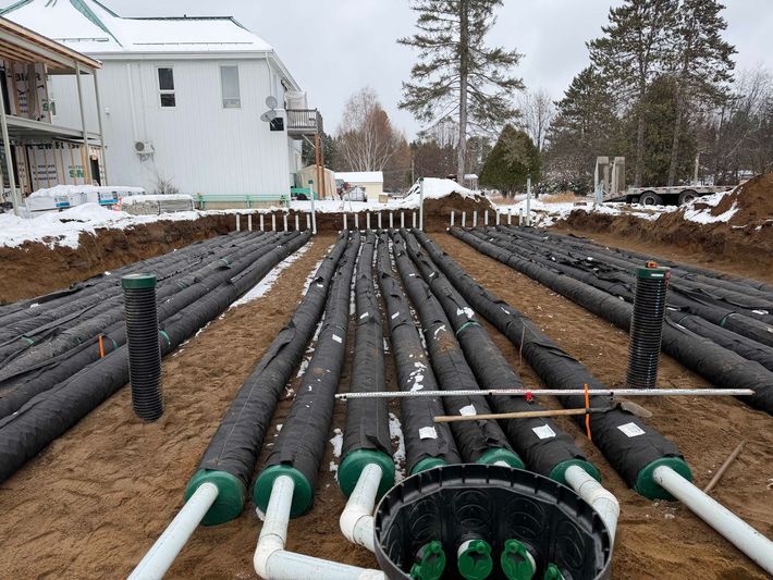 Des rangées de tuyaux de drainage noirs, recouverts de tissu, sont installées dans une tranchée sur un chantier de construction enneigé.