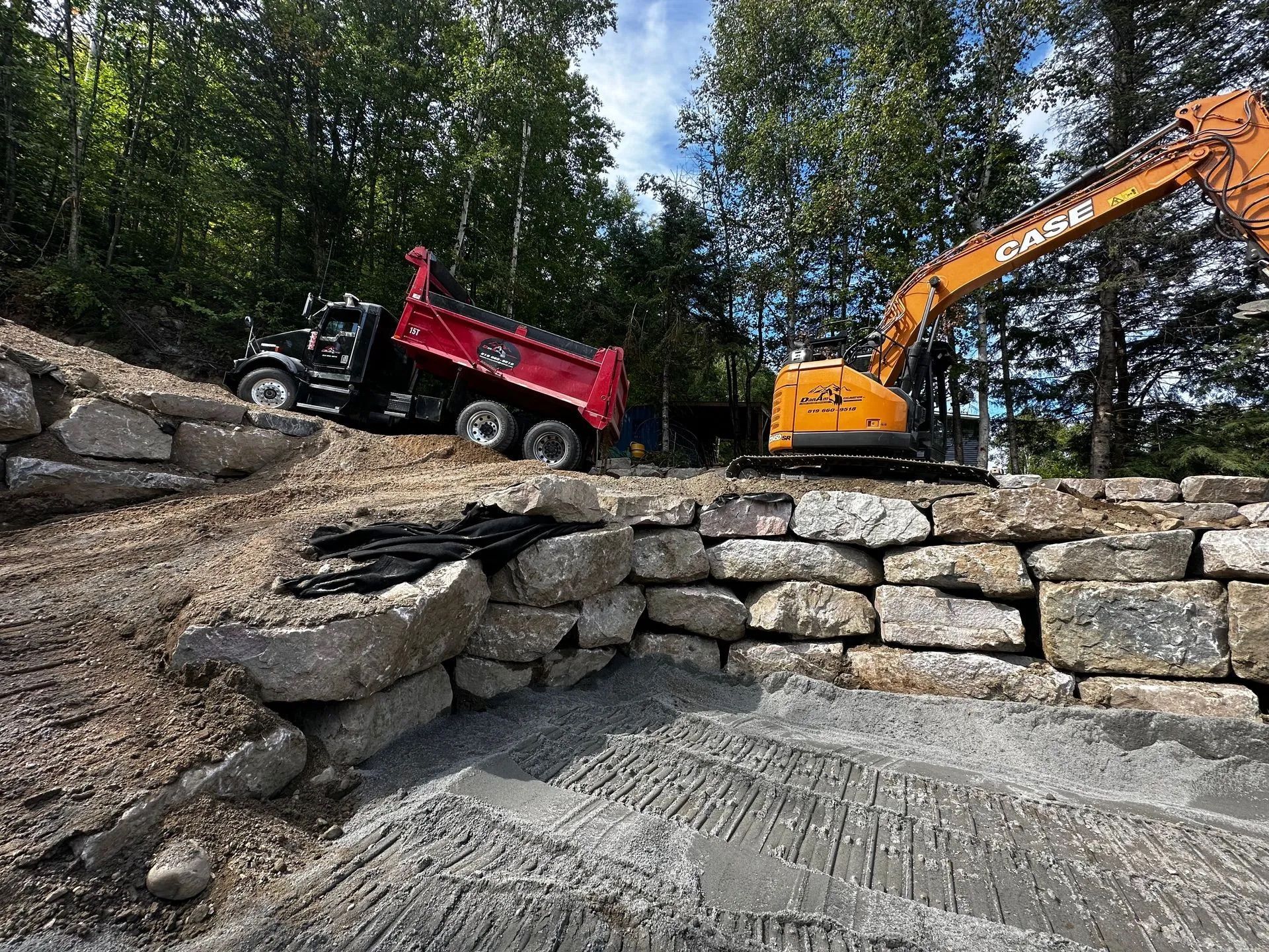 Un camion-benne rouge et une excavatrice jaune sont stationnés sur un terrain en pierre en terrasses, en cours d'aménagement dans une zone boisée.
