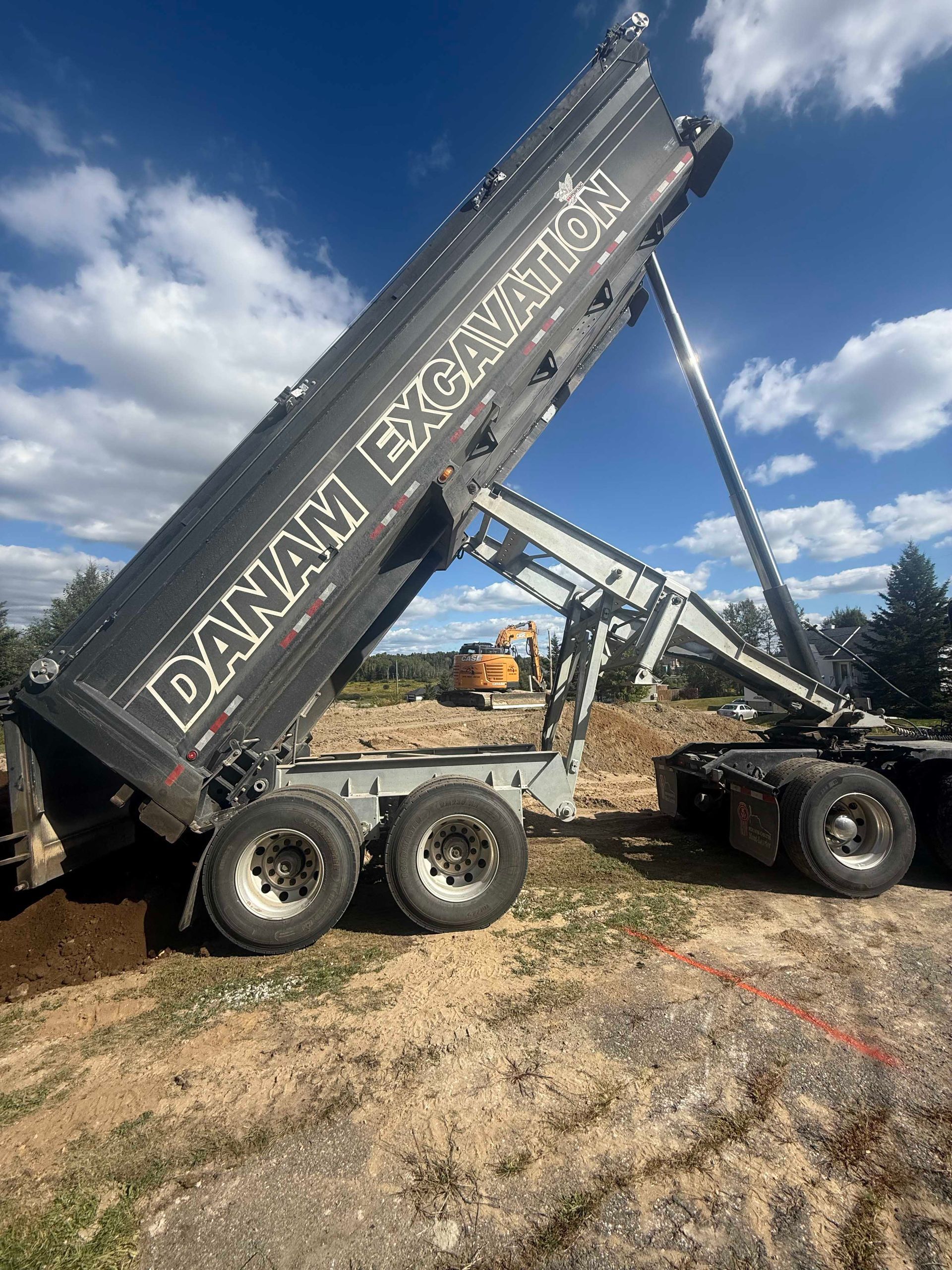 Un camion-benne noir de la société Danam Excavation est stationné sur un terrain vague, sa remorque étant entièrement levée pour décharger de la terre.