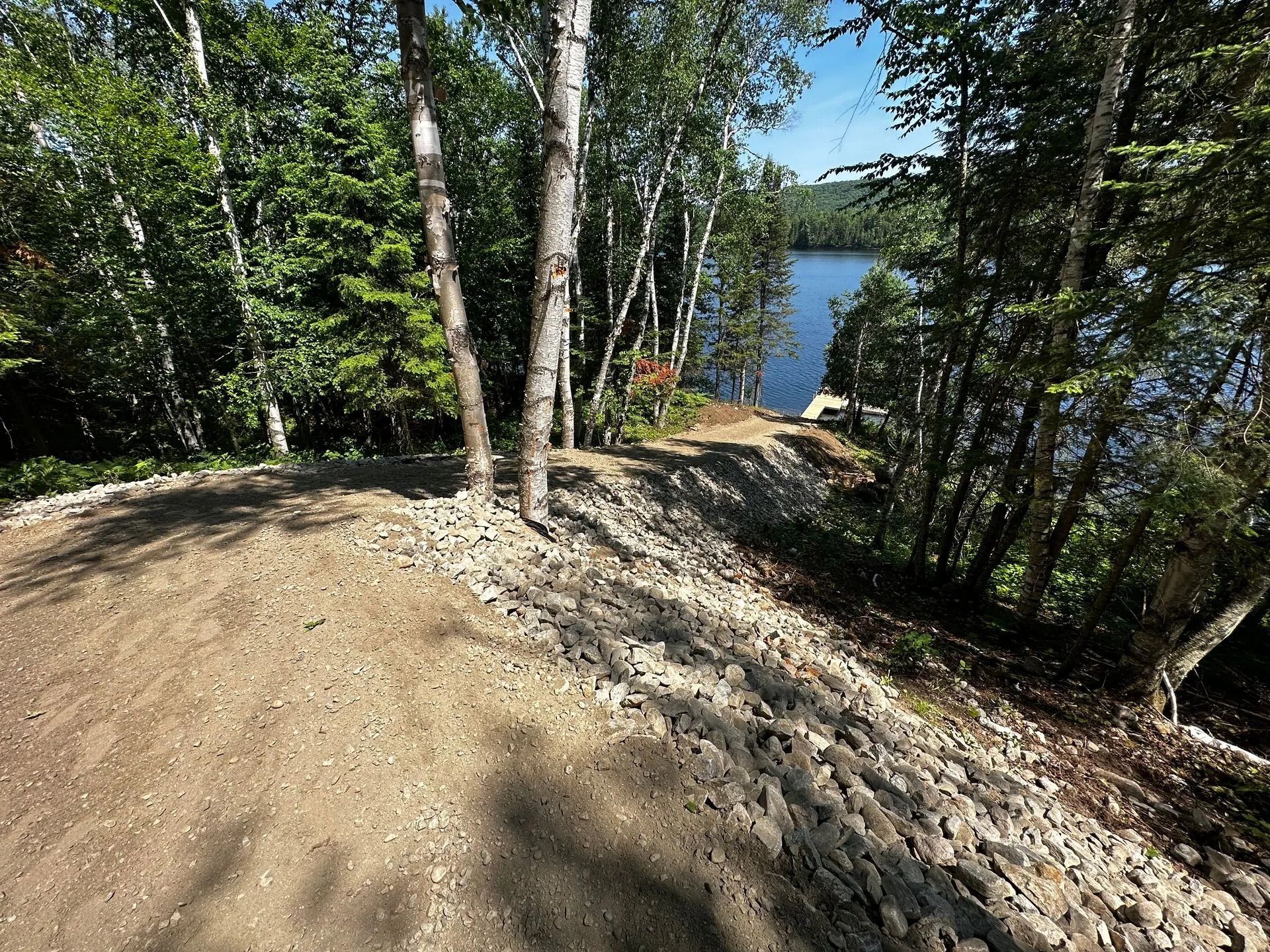 Un chemin de terre descend un versant boisé vers un petit ponton en bois sur un lac calme, sous un ciel d'un bleu limpide.