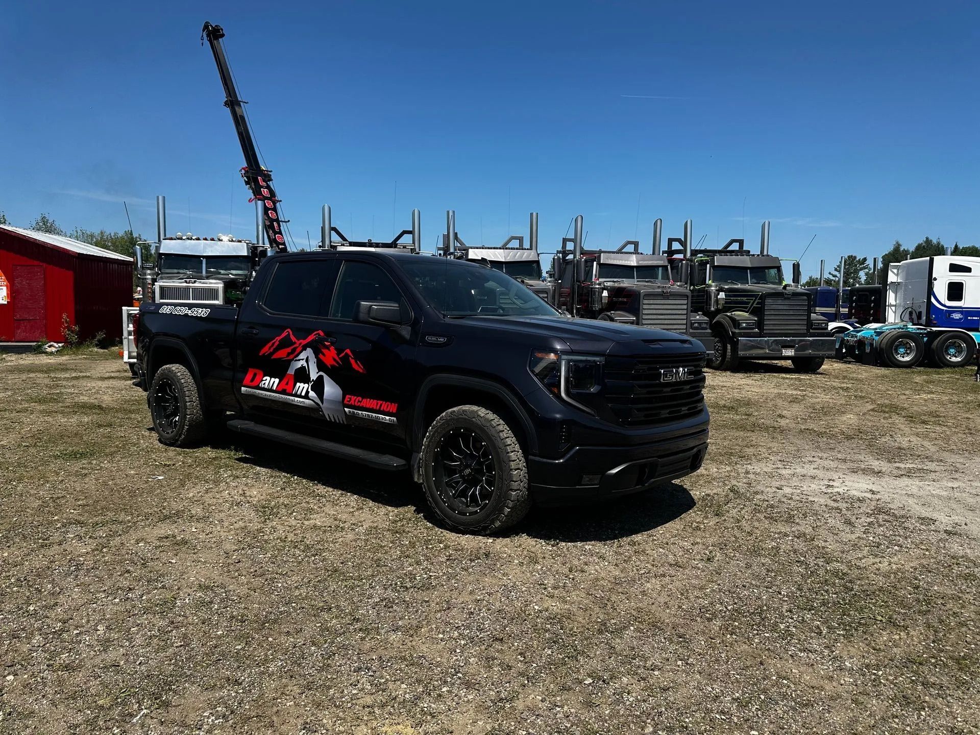 Un pick-up GMC noir est garé sur un terrain gravillonné, avec de gros semi-remorques et une grue visibles en arrière-plan.