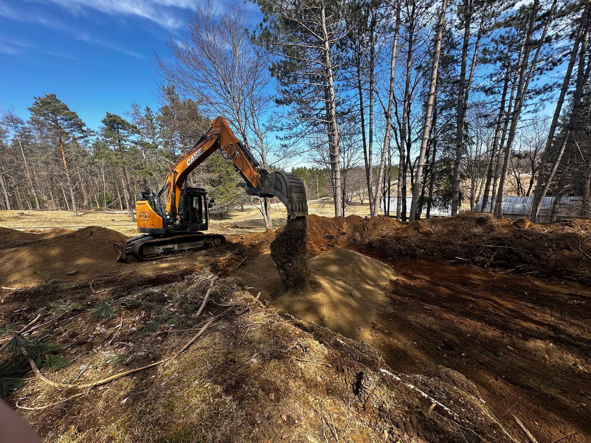 Une pelleteuse jaune creuse un terrain vague en pleine terre, entouré d'arbres dénudés, sous un ciel d'un bleu éclatant.