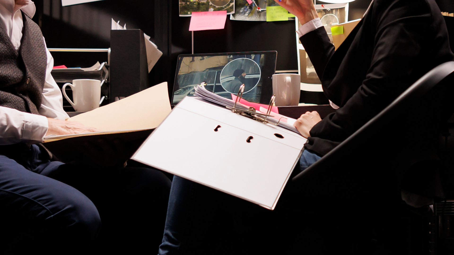 Two people in an office. One holds a file, the other gestures. A laptop and a cup sit on the desk.