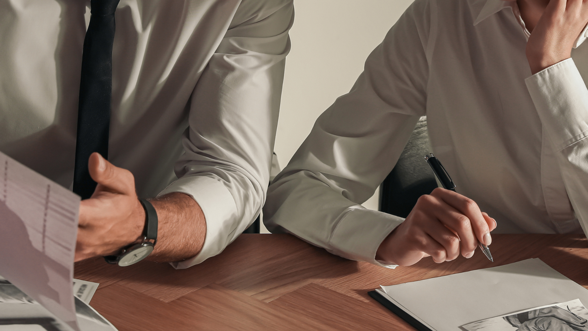 Two people in white shirts at a desk, reviewing documents. One holds papers, the other writes notes.