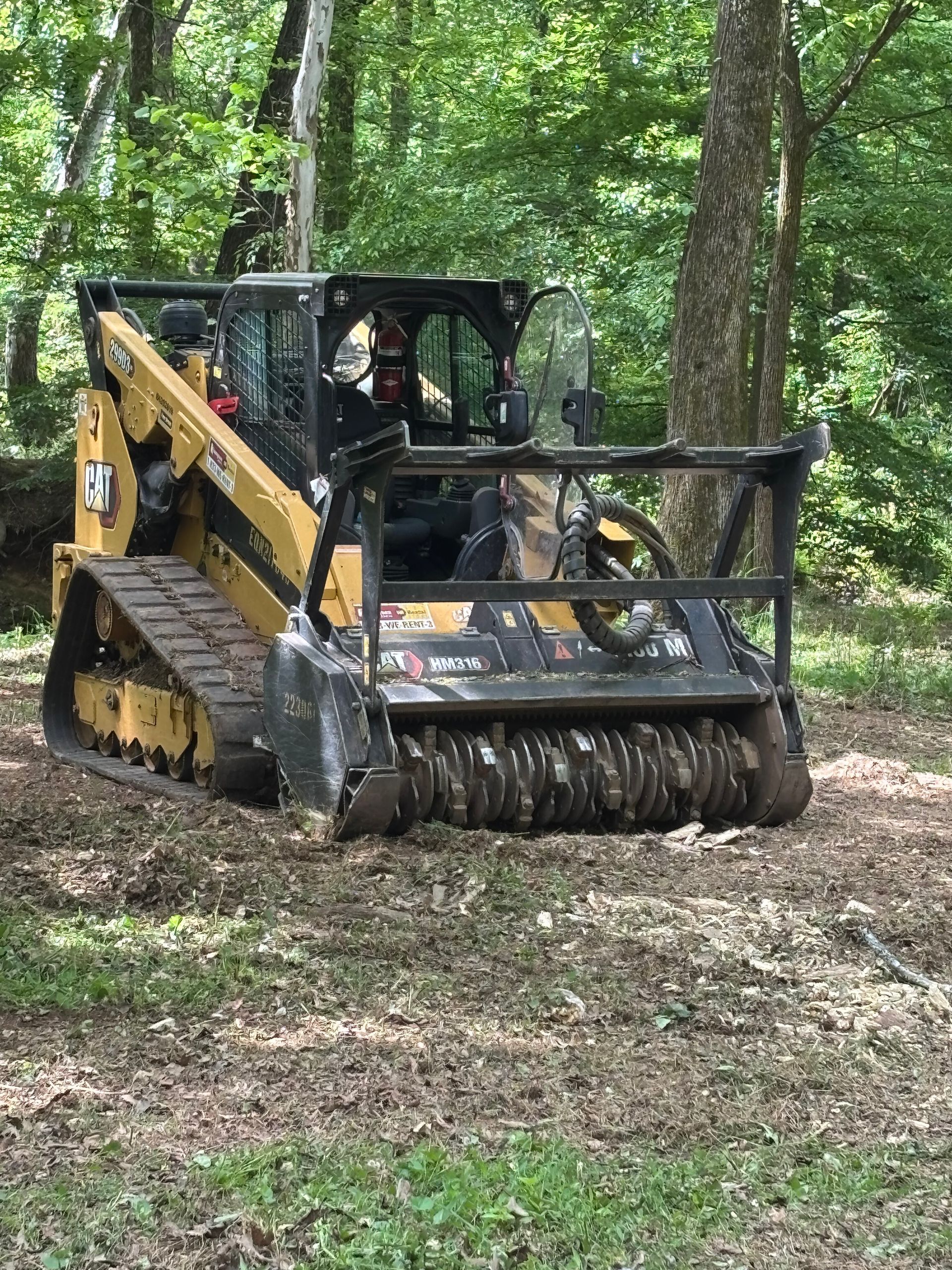 Yellow skid-steer clearing leaves in a wooded area with a brush cutter attachment