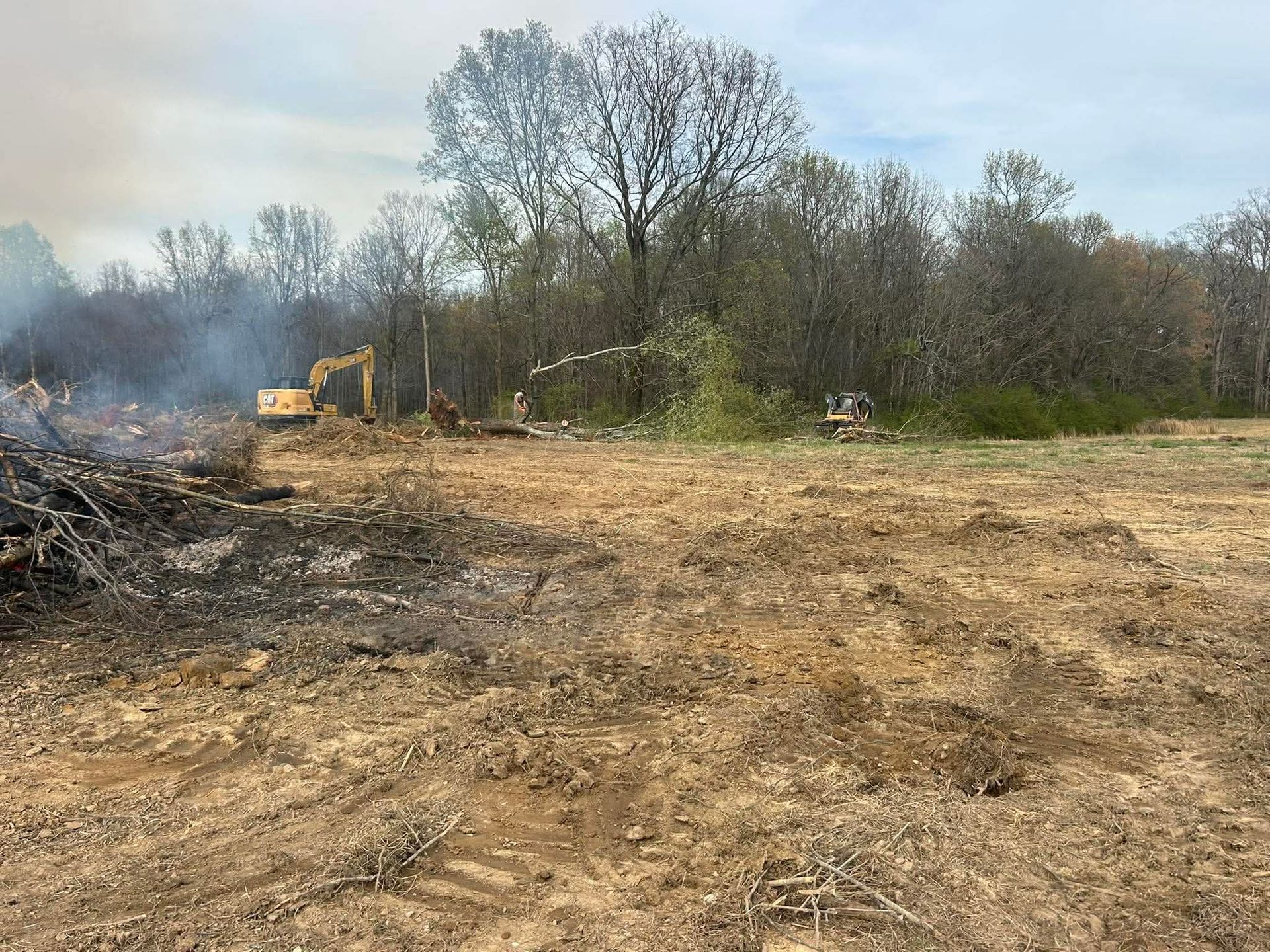 Cleared dirt lot with burn piles and a yellow excavator near trees under a hazy sky