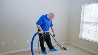 Man vacuuming beige carpet in an empty room with a blue hose and a window on the right