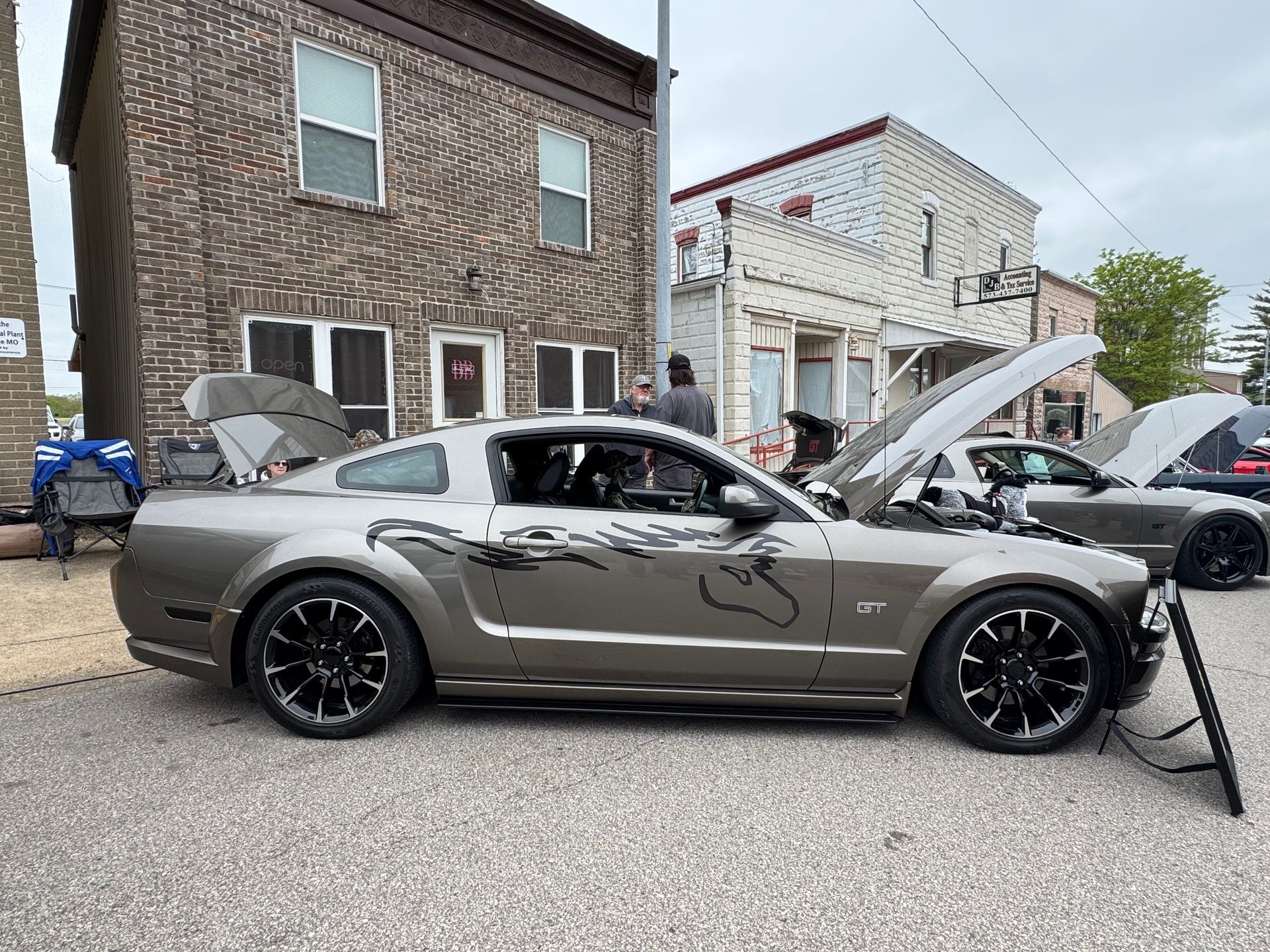 Gray Ford Mustang with hood open at an outdoor car meet beside brick buildings