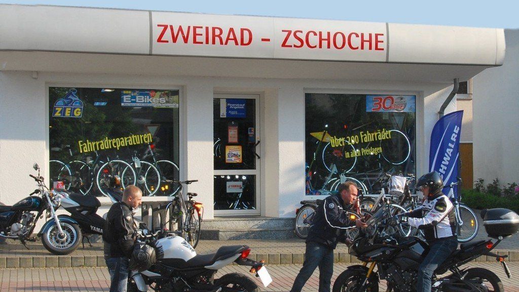 Motorradfahrer versammeln sich vor dem Fahrradgeschäft Zweirad-Zschoche, das große Schaufenster mit Fahrradaufklebern hat.