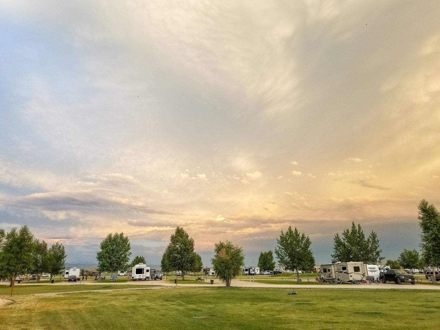 Camping ground with RVs under a cloudy sky at sunset; green grass, trees, and colorful clouds.