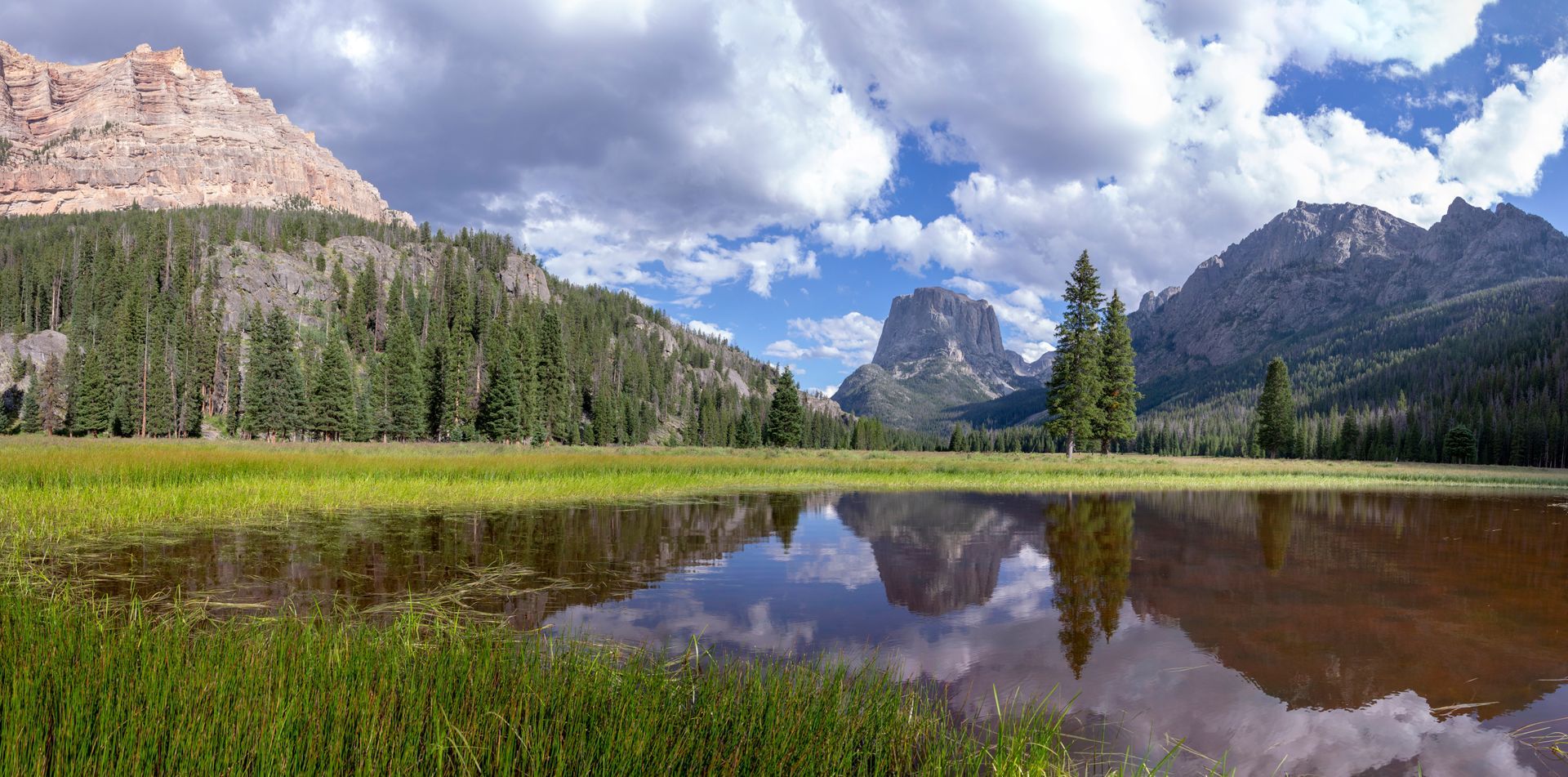 Mountain landscape reflected in calm water. Green grass in the foreground. Blue sky with clouds.