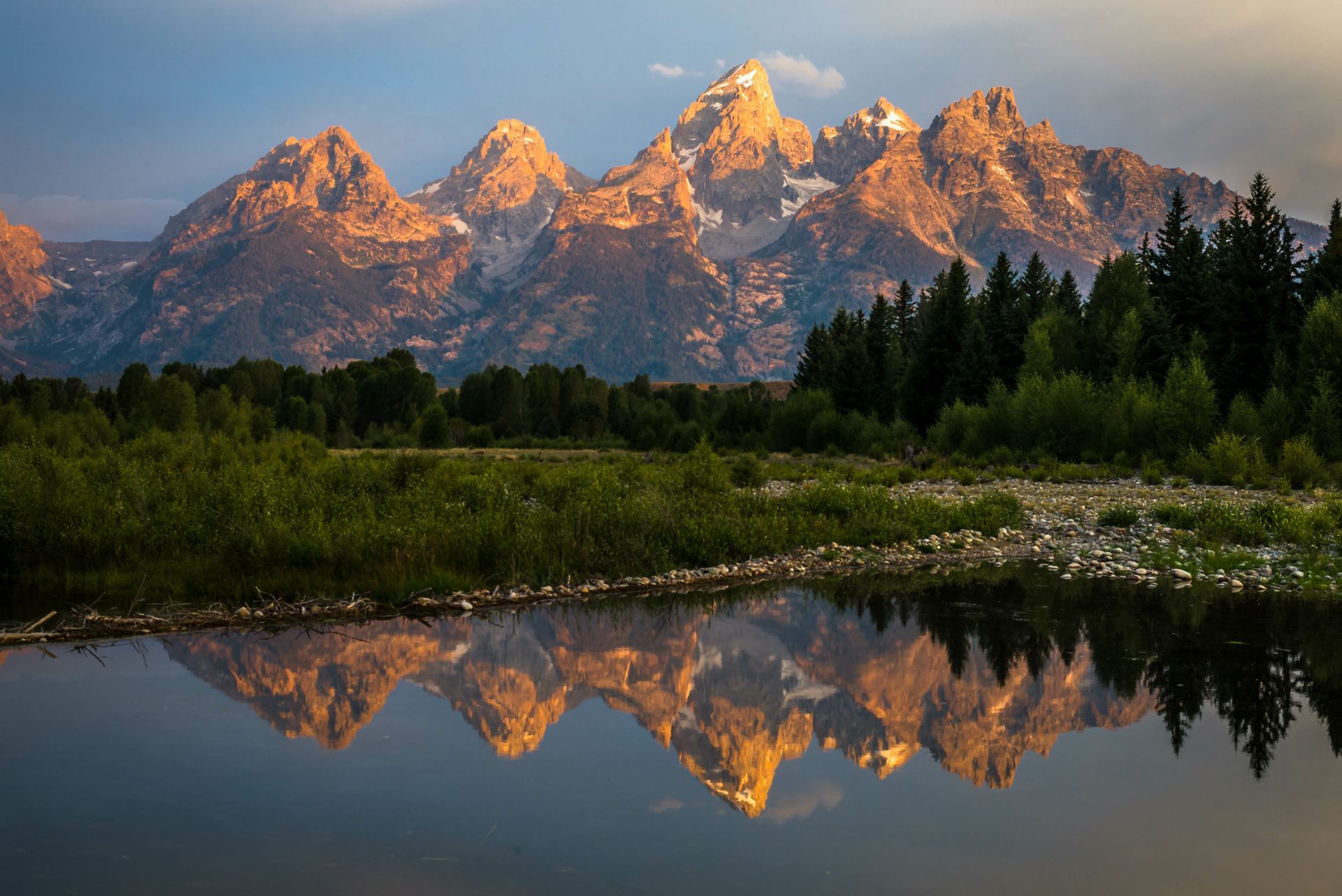 Mountains reflected in water, lit by golden sunlight; trees in foreground.