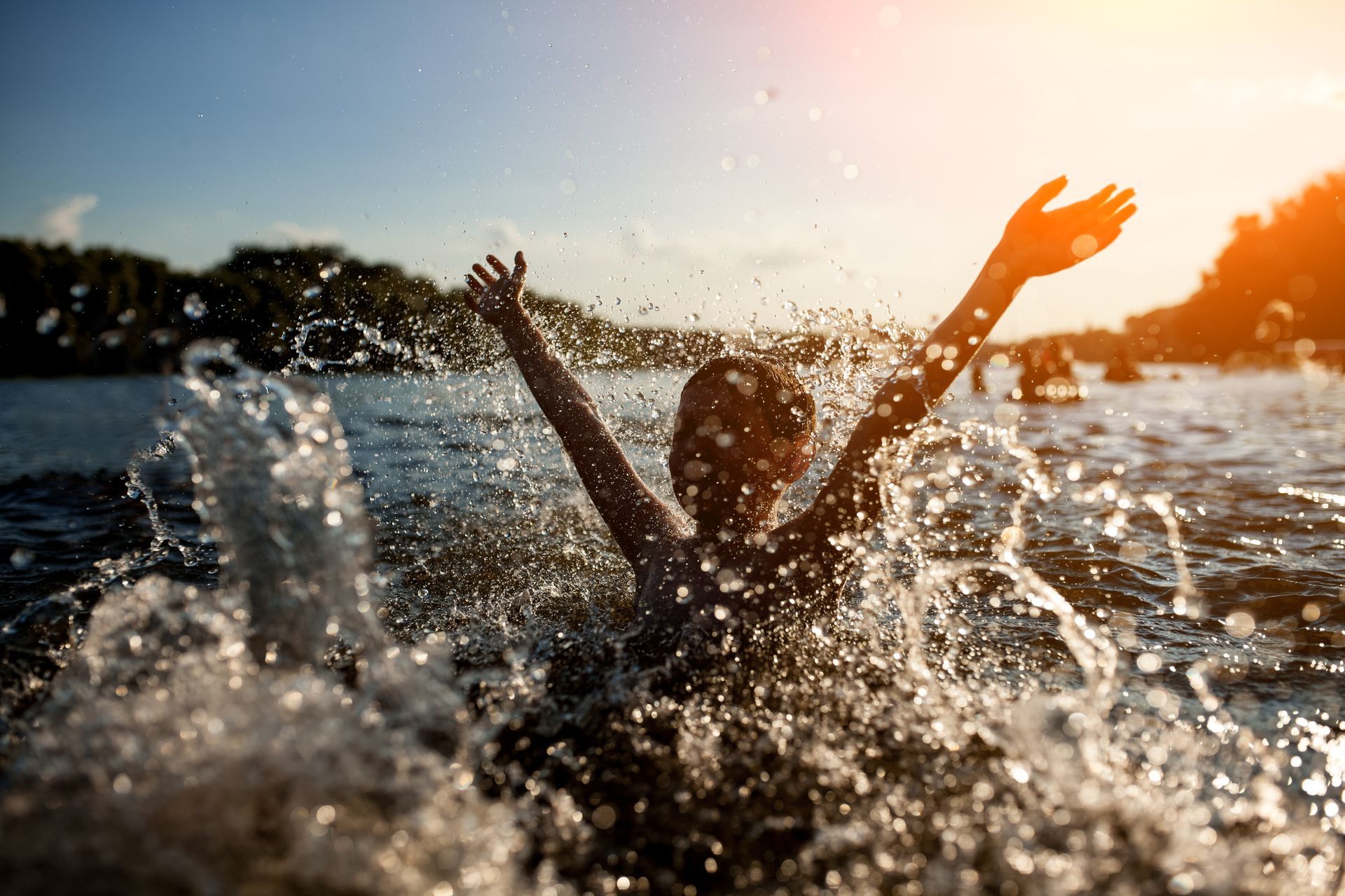 Person in water with arms raised, splashing. Sun shining, trees in background.