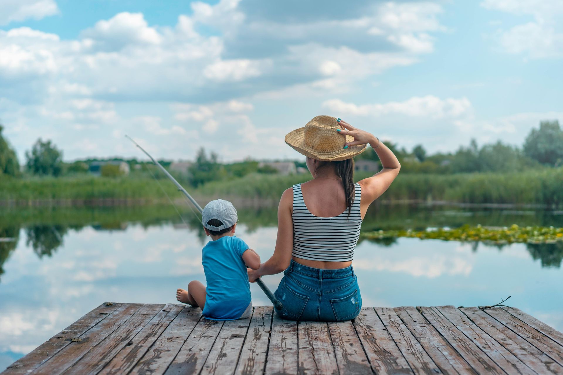 Woman and child fishing together on a wooden dock overlooking a serene lake, under a cloudy sky.