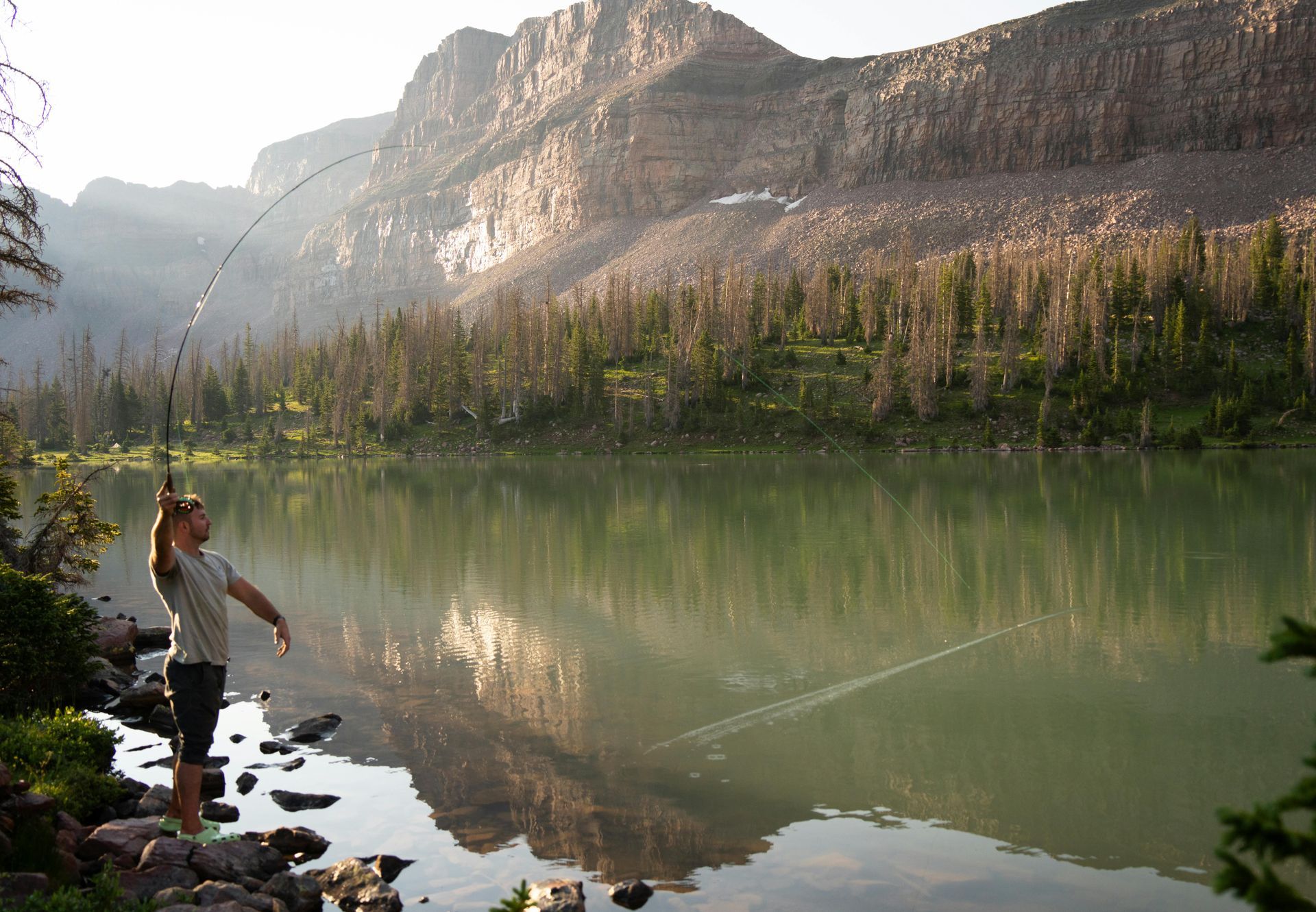 Man fly-fishing in a mountain lake, with a rocky shore and forested mountains in the background.