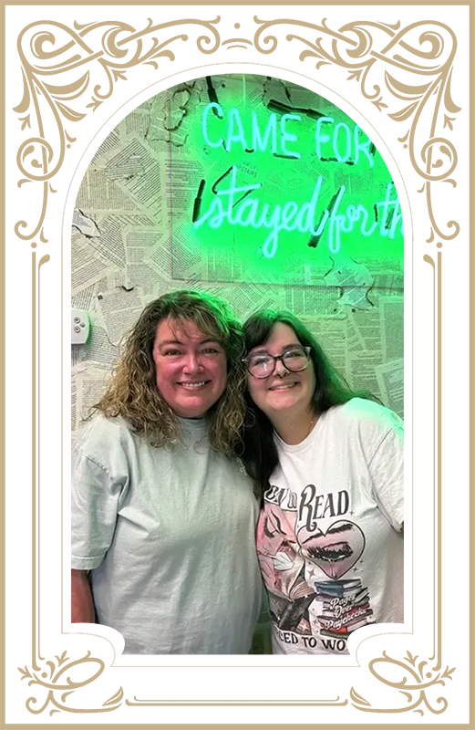 Two smiling friends posing in front of a neon green sign and ornate white frame border