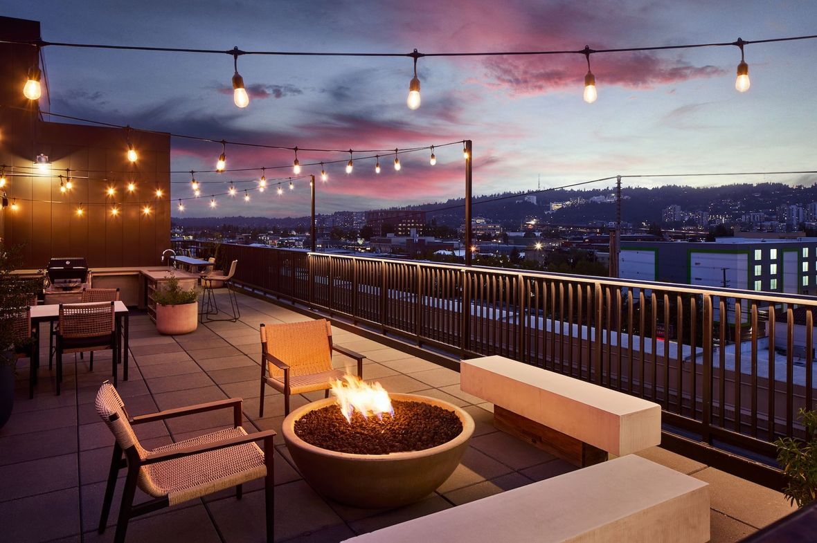 Rooftop lounge at dusk with string lights, fire pit, and city skyline view