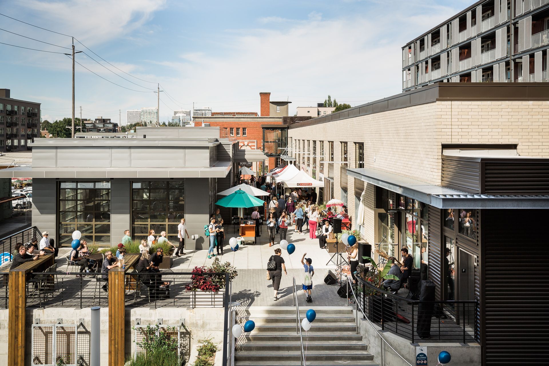 Crowded outdoor plaza with steps, shops, and people walking under sunny skies