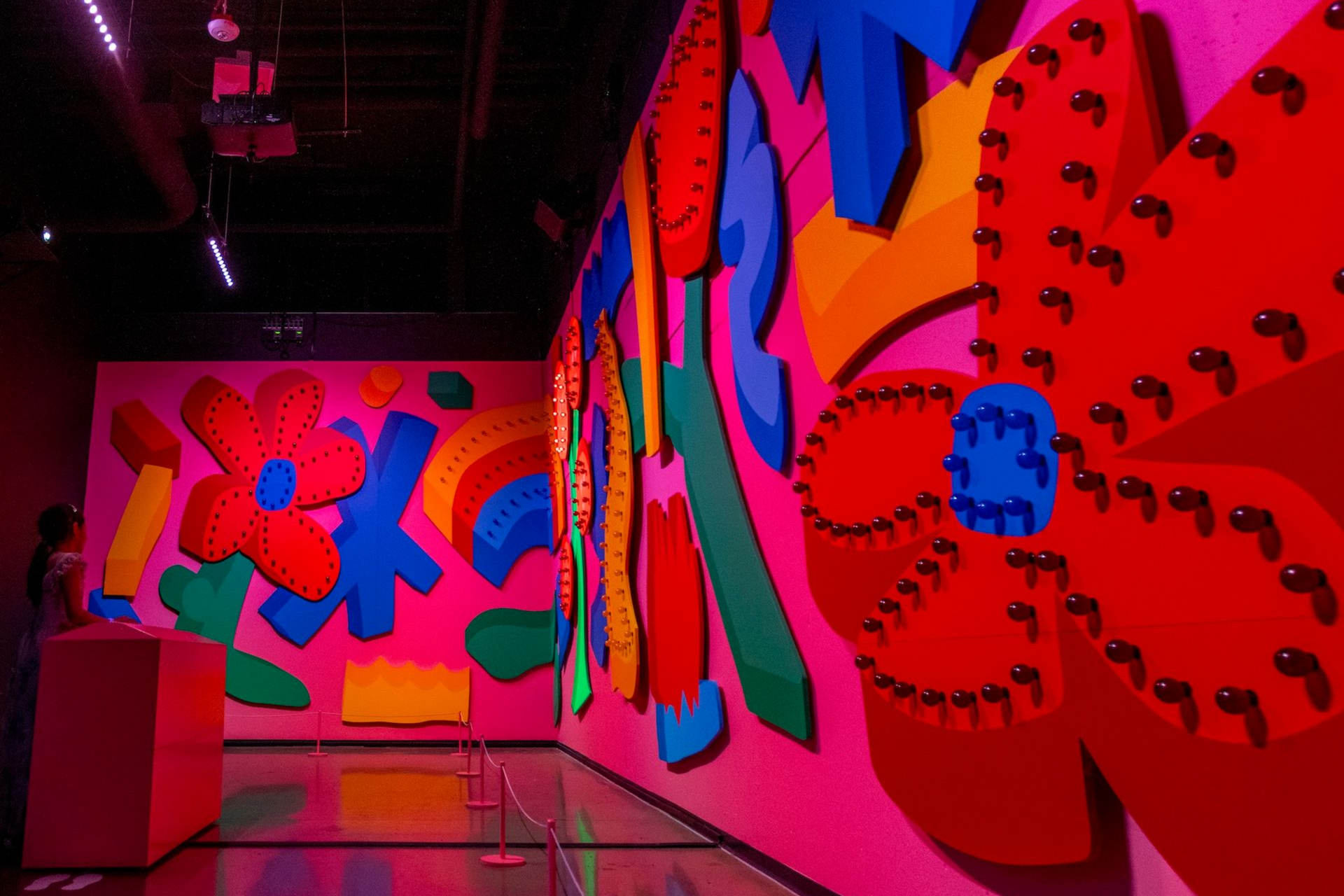 Neon-lit hallway with bright pink wall art featuring large colorful floral patterns and a reflective floor