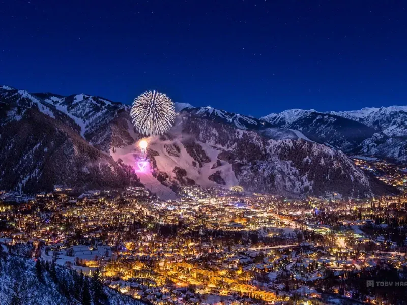 Fireworks over a snowy mountain town at night, illuminating the buildings and surrounding landscape.
